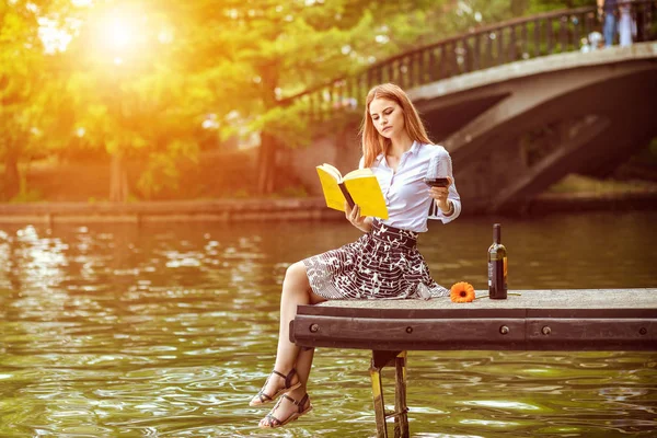 Şarap - Modern yaşam tarzı ve Özgürlük kavramı ile bin yıllık kız zevk gün Park - sıcak güneş renk filtresi sesi içme cam elinde kitapla dock parkta oturan genç hipster kadın