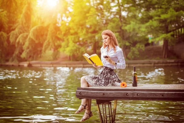 Genç hipster rahatlatıcı kadın üstünde rıhtım park okuma bin yıllık kız gün park, sıcak güneş renk filtresi sesi zevk ile içme bardak kırmızı şarap, Modern yaşam tarzı ve Özgürlük kavramı kitap