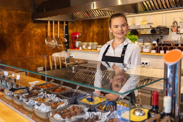 Smiling waitress wearing black apron standing behind counter in small family fast food restaurant looking at camera. Young woman serving food, Small business and entrepreneur concept.