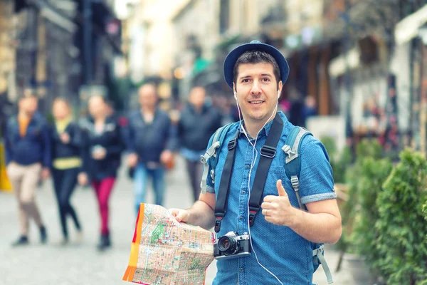 Reisender mit trendigem Blick, Landkarte mit Daumen hoch auf Straße voller Restaurants und Geschäfte der europäischen Stadt, junger Mann genießt Stadtatmosphäre beim Fotografieren mit Fotokamera Touristen die Welt erkunden — Stockfoto