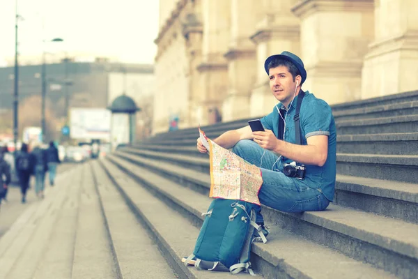 Smiling caucasian man using mobile phone while sitting on steps, happy tourist searching for map directions on smartphone, traveler with backpack and hat looking at city attractions