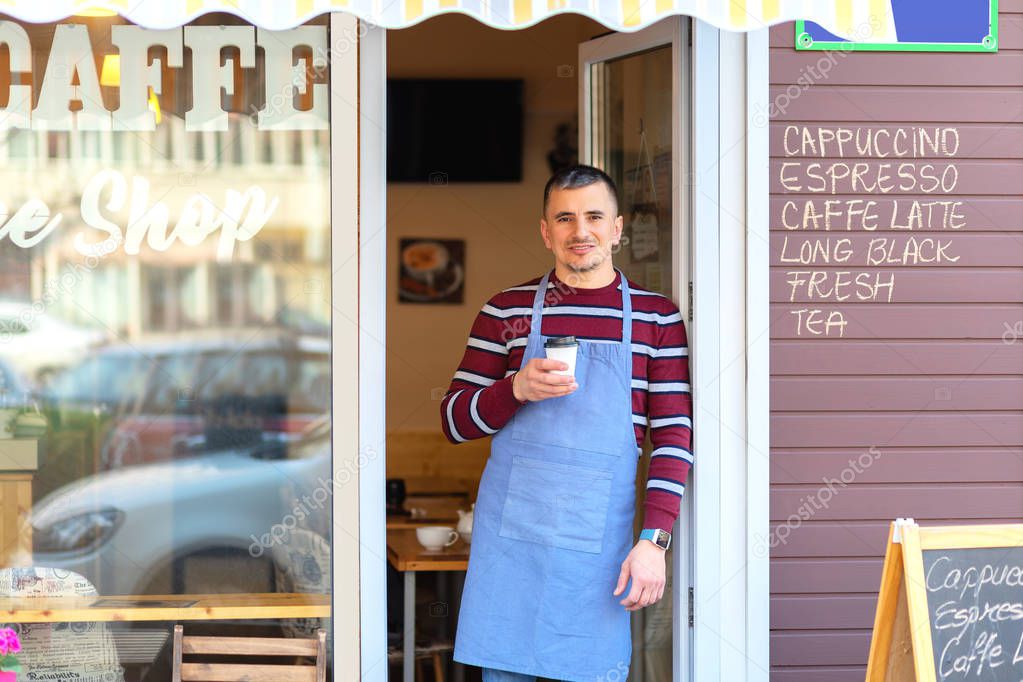 Happy barista standing at cafe entrance, portrait of mature business man attend new customers in her coffee shop, happy male owner holding cup of take-away cofee