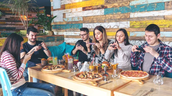 Social media addiction concept with young people photographing food in rustic restaurant, happy friends taking picture of pizza and hamburgers with mobile phones to post online, connecting millennials