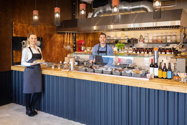 Yemek servisi yapan güler yüzlü garsonlar, açık mutfaklı küçük restoranda mutlu işletme sahipleri ile modern bistroda karşı servis, yemek tadımı için içerideki insanları davet ediyor, catering hizmeti