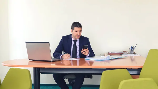 Akıllı telefon üzerinde yazan ve ofiste masada otururken bir belge yazma emin iş adamı, online borsa istatistikleri görüntülemek için bir cep telefonu kullanarak resmi giysiler içinde Olgun işadamı