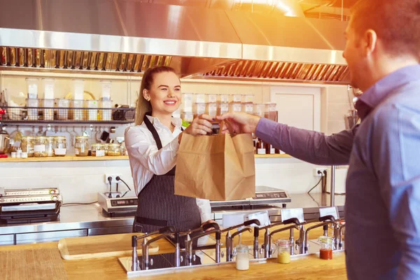 Restoranda tezgahta müşteriye hizmet veren apron giyen neşeli garson - Küçük işletme ve hizmet konsepti genç kadın sahibi ile çevre dostu kağıt torba veren online müşteriye yiyecek götürmek