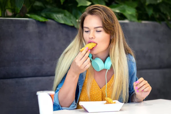 Fast food'da çıtır çıtır şeritler yiyen genç gülümseyen trendy kadın - Mutlu moda hipster kız rahat giyim ve kızarmış tavuk tutarak kulaklık giyen ve restoran eatery de sağlıksız gıda tatma
