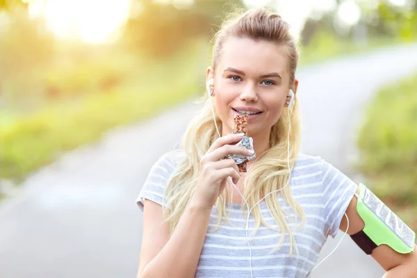 Athletic woman eating protein bar after jogging in park - closeup face of young sporty girl resting while taking a bite a nutritive fruit bar - beautiful fitness jogger eating energy snack outdoor