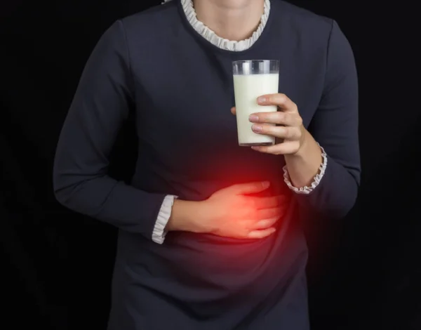 La niña se agarra al estómago y sostiene un vaso de leche, un fondo negro, dolor y acidez estomacal — Foto de Stock