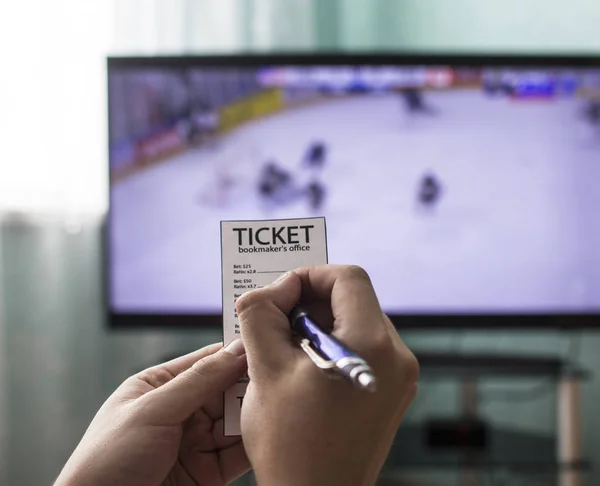 Человек держит букмекер билет и ставки на спорт, по телевизору идет хоккей, крупный план — стоковое фото