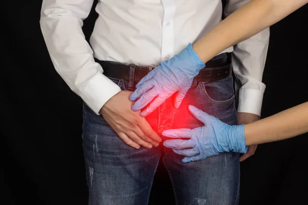 En man i jeans och en vit skjorta håller på ljumsken, kvinnors händer i medicinska handskar håller en man av ljumsken. svart bakgrund, prostatit — Stockfoto