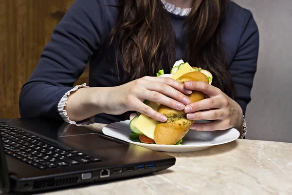 La chica tiene un sándwich en el lugar de trabajo detrás de la computadora portátil — Foto de Stock