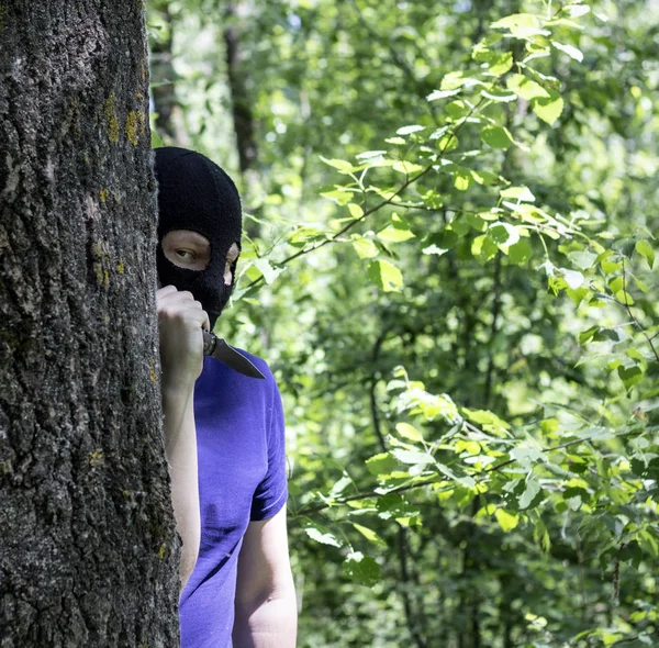 Maniac, the killer in a black mask is behind a tree with a knife, close-up, killer with a knife in the open air