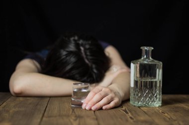 Sarhoş bir kız kafasını masaya, onun elinde bir bardak alkol ve bir şişe, ahşap bir masa, siyah bir arka plan koydu