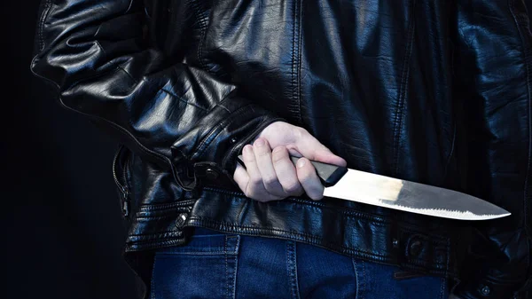 Un hombre con una chaqueta de cuero sostiene un cuchillo detrás de su espalda, un peligro de fondo negro —  Fotos de Stock