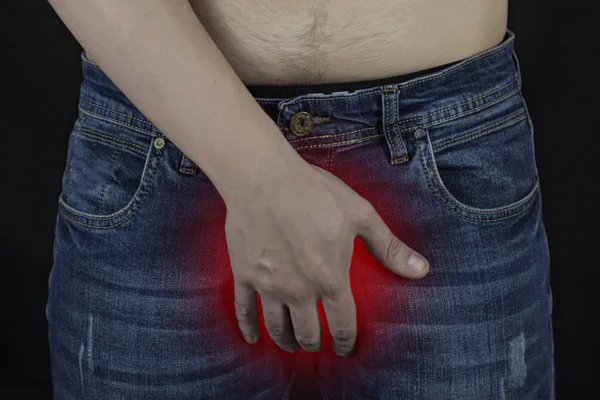 Un hombre en jeans sostiene sus manos en la ingle, un fondo negro, una ingle roja — Foto de Stock