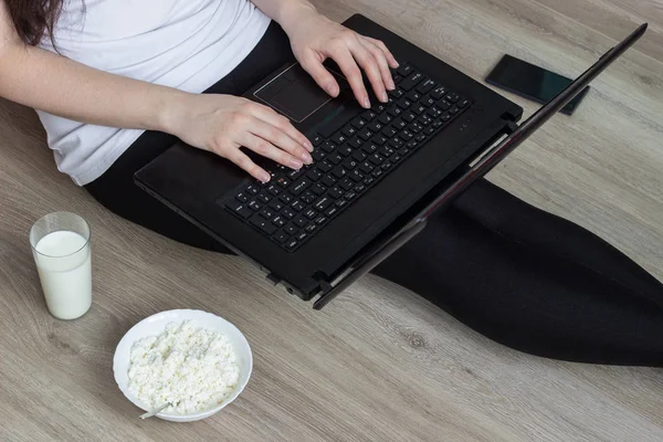 Chica que trabaja detrás de un ordenador portátil, teléfono, vaso de leche y un plato con queso cottage — Foto de Stock