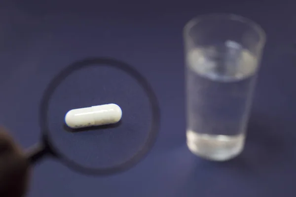 Tabletki pod powiększenie lupy, szklankę wody na niebieskim tle — Zdjęcie stockowe