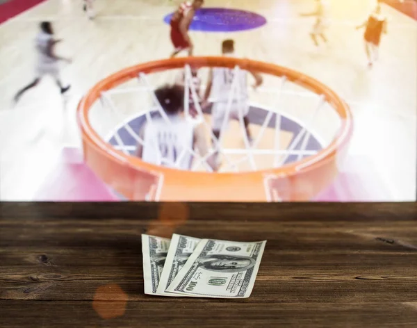 Dinero dólares en un fondo de madera en el fondo de un televisor que muestra el baloncesto — Foto de Stock