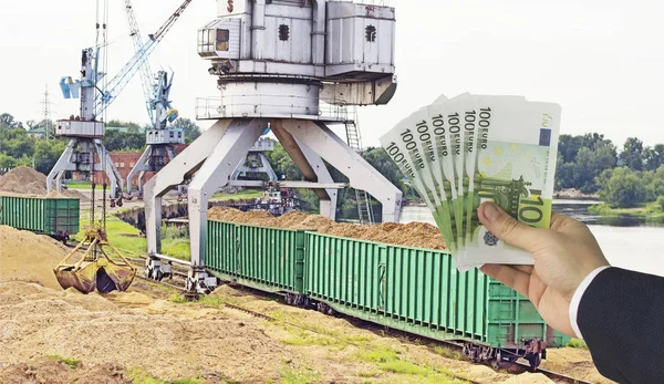 Une main avec de l'argent en euros contre une grue portuaire rend le chargement de copeaux de bois dans des wagons de fret, l'exportation en euros — Photo