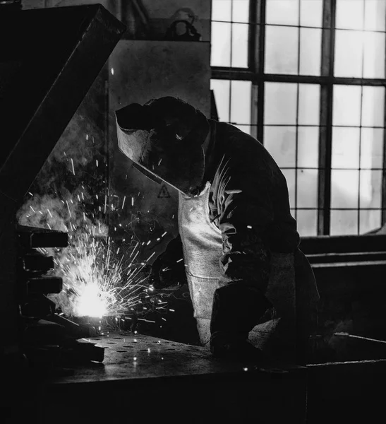 Soudeur à l'usine dans un masque de soudage soude des pièces métalliques, soudage et étincelles, noir et blanc, support de soudage — Photo