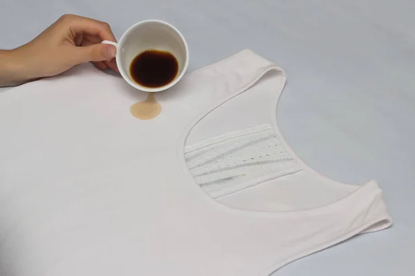 Дівчина п'є чашку кави на білій майці, борючись з плямами на одязі, перевіряючи засоби для видалення плям, каву та плями, крупним планом, рукою, чашкою — стокове фото