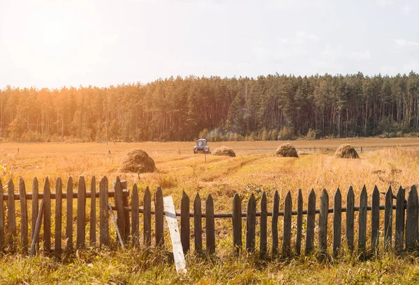 Een oude trekker plows een veld in een dorp, hooibergen en een oude hek op het veld staan, er is een bos aan de horizon, machines — Stockfoto