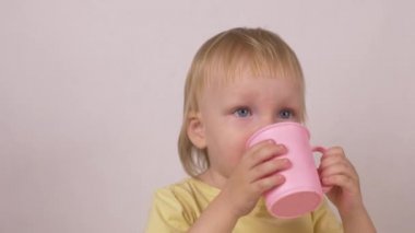 Küçük sarışın beyaz kız pembe bir kupa biraz su içer ve görünüyor kamera, beyaz arka plan, yakın çekim, geleneksel