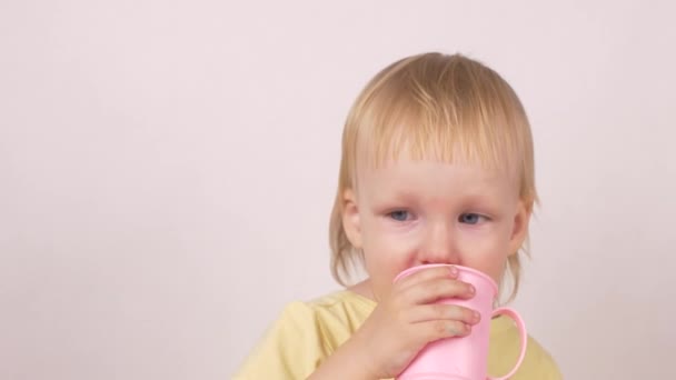 Pequena menina branca loira bebe um pouco de água de uma caneca rosa e olha para a câmera, fundo branco, close-up — Vídeo de Stock