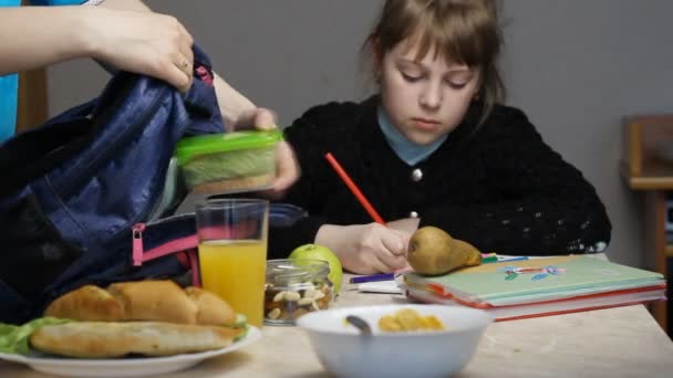 Мама собирает обед в школу, кладет контейнер с едой в школьную сумку, девочка делает домашнее задание — стоковое видео