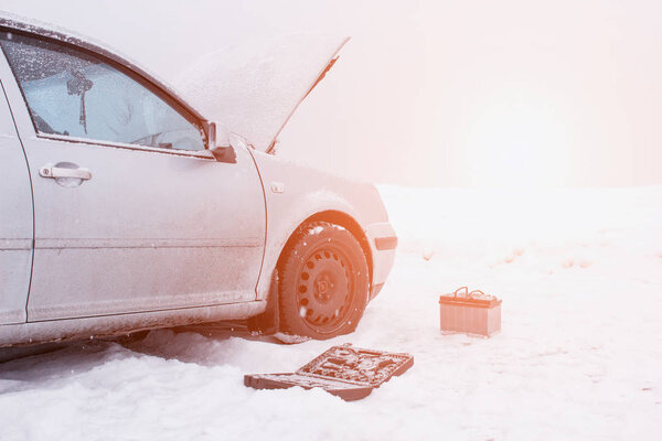 A car with a raised hood on a background of a snowy field, tools and a discharged car battery near the car, the concept of a car breakdown in winter