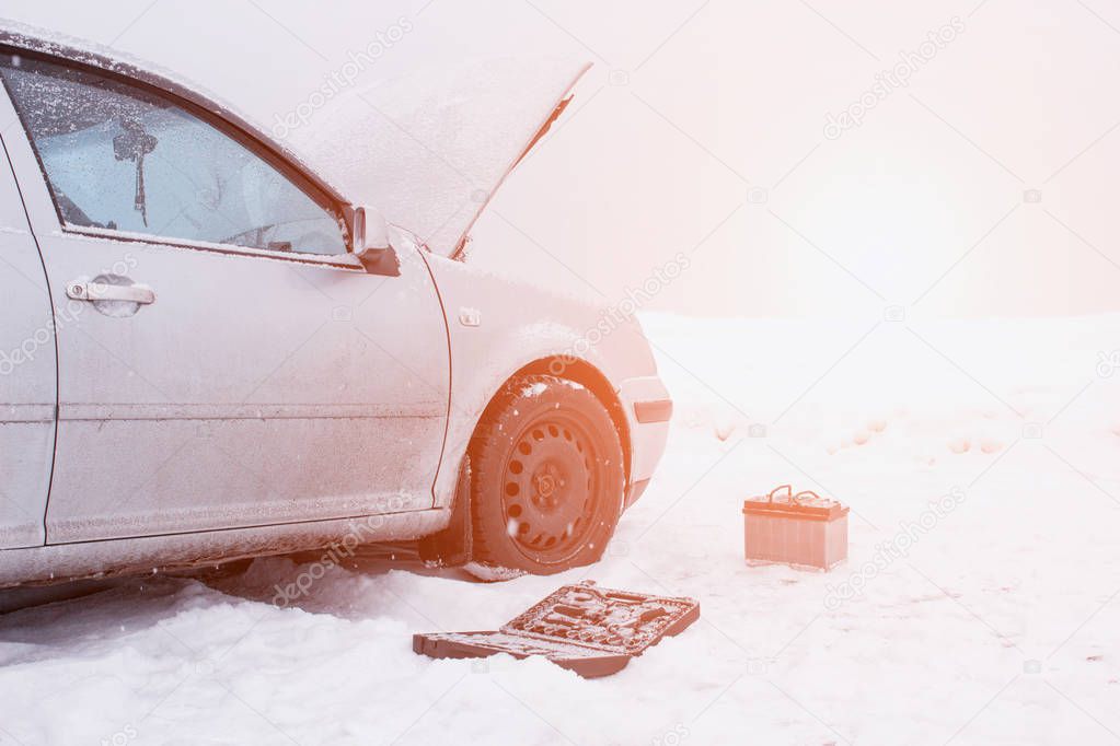 A car with a raised hood on a background of a snowy field, tools and a discharged car battery near the car, the concept of a car breakdown in winter