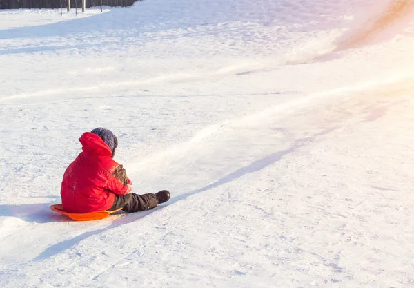 Parlak kırmızı elbiseli küçük çocuk soğuk kış havasında tepeden aşağı yuvarlanıyor. — Stok fotoğraf
