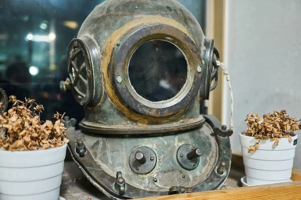 Vintage diving helmet with rusty metal displayed on a shelf along with dry flowers on both sides.