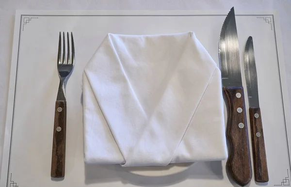 A silverware and napkin on a white plate neatly displayed on white sheet of paper on a table.