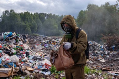 Gaz maskeli bir adam çanta içeriğini denetler. Çöplükte çalışıyor. Çevresinde yakar ve plastik çöp dumanlar