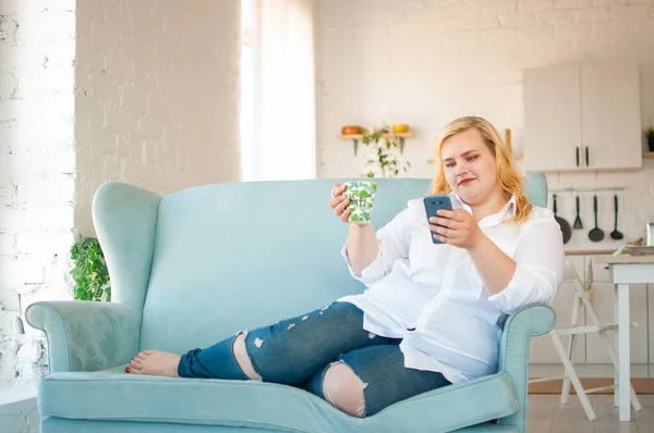 Beautiful plus size girl sits on the couch and communicates using a smartphone — Stock Photo, Image