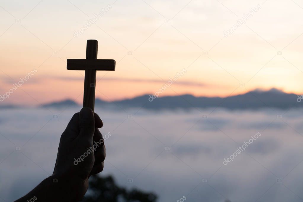 Woman hand holding holy lift of christian cross with light sunset background.