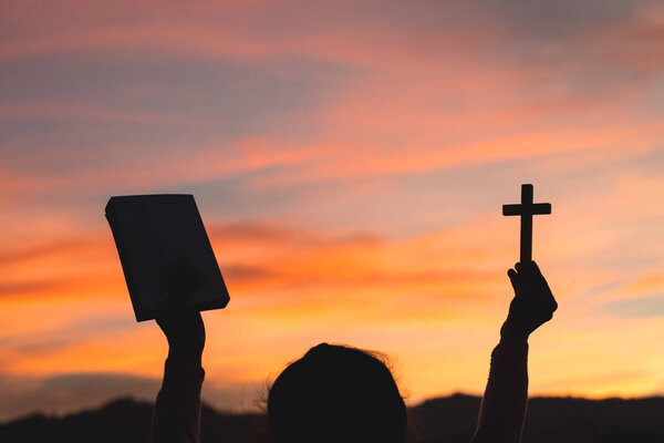 Silhouette of Young woman hands holding holy Bible and lift of christian cross, religion symbol in light and landscape over a sunrise, background, religious, faith concept 
