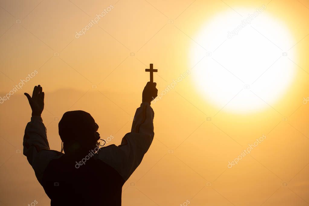 Silhouette of a man praying with a cross in hand at sunrise,  re