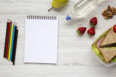 Sağlıklı okul öğle yemeği kutusu beyaz ahşap zemin üzerinde defter ve renkli kalemler ile düz yatıyordu. Yukarıdan. Üstten Görünüm. Metin için yer.