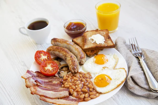 Full english breakfast with fried eggs, bacon, sausages, beans and toasts on white wooden background. Closeup. Side view.