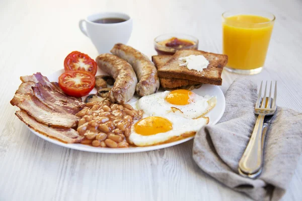 Full english breakfast with fried eggs, bacon, sausages, beans and toasts on white wooden background. Closeup. Side view.