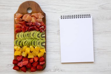 Üstten görünüm, doğranmış taze meyve kesme tahtası ve defter Beyaz ahşap zemin üzerine düzenlenmiştir. Düz yatıyordu.