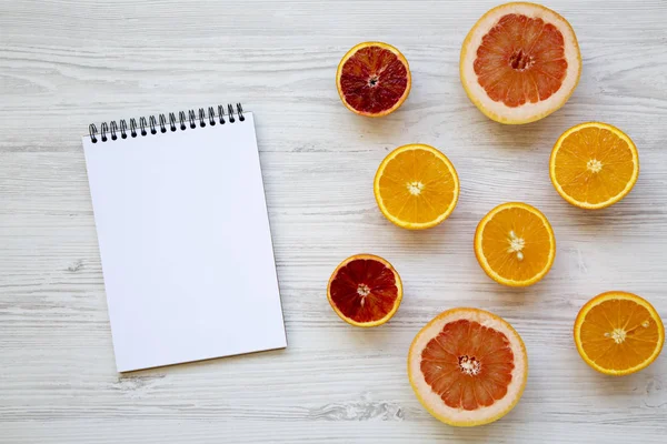 Citrus fruits (orange, grapefruit, sicilian orange) with blank notepad on white wooden background, from above. Flat lay. Top view.