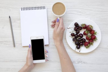 Smartphone, latte, defter, çilek ve kiraz Beyaz ahşap arka plan, üstten görünüm üzerinde eller. Düz yatıyordu. Yukarıdan.