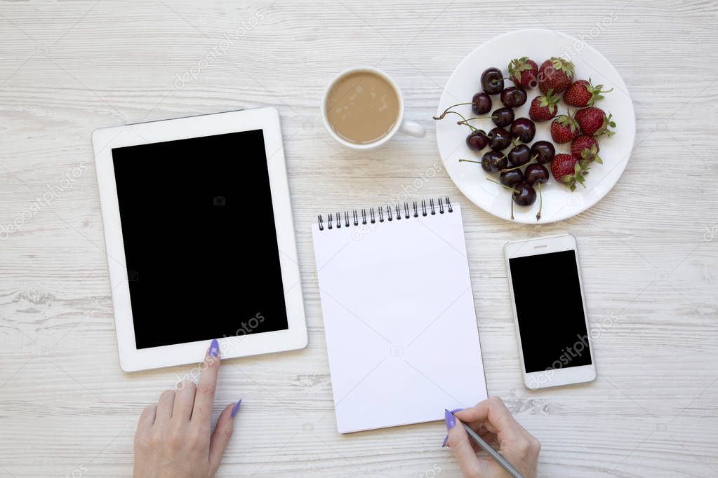 Top view, modern feminine workspace with female hands, smartphone, tablet, notepad, berries and latte on white wooden background. Flat lay. From above.