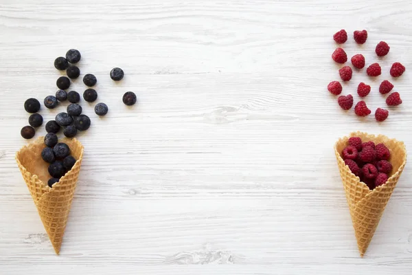 Waffle sweet ice cream cones with raspberries and blueberries over white wooden background, top view. Flat lay. Copy space.