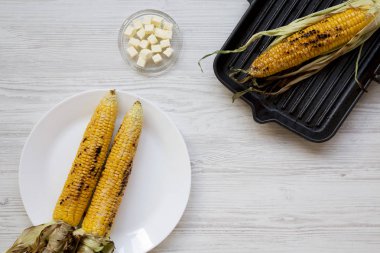Izgara tava ve beyaz tabak, tereyağı, üstten görünüm üzerinde ızgara mısır koçanı. Yukarıda, ek yükü.