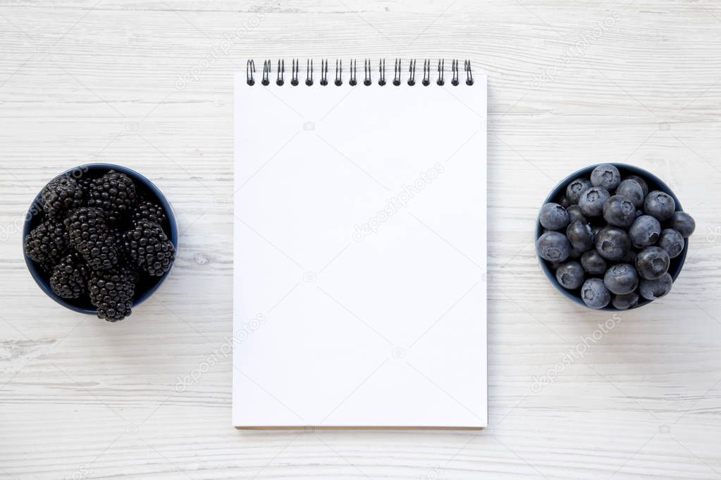 Full bowls of berries with blank notebook on a white wooden background, top view. From above, overhead, flat lay.